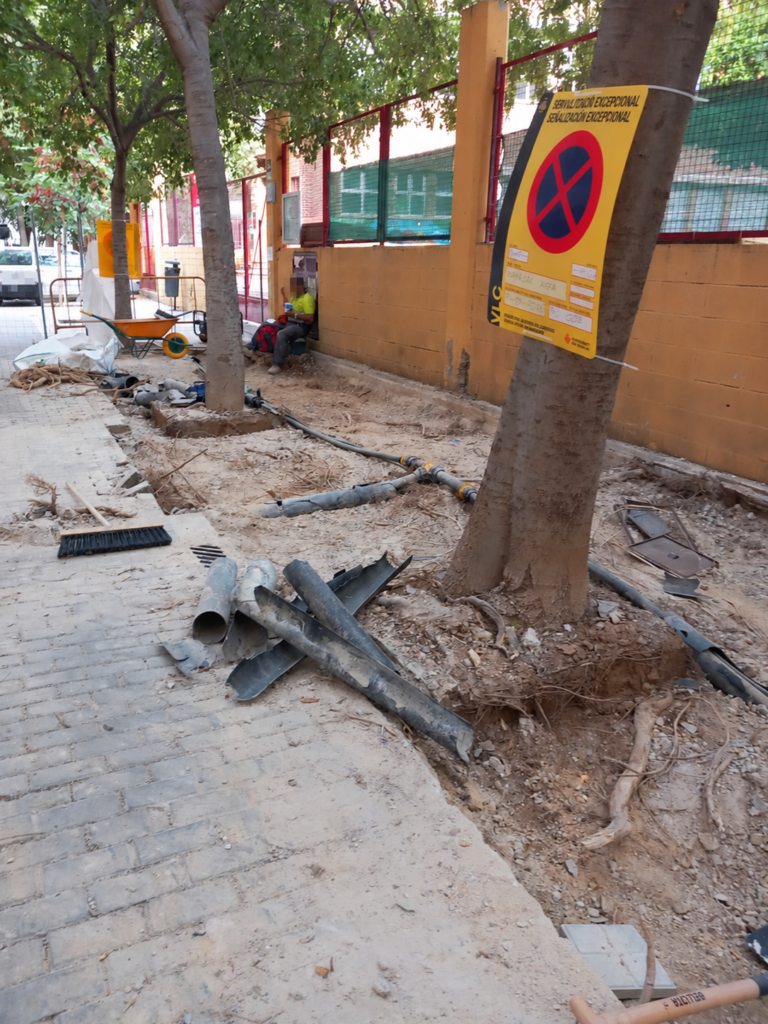 Obra de reparación de una acera con raíces expuestas y servicios urbanos visibles.
